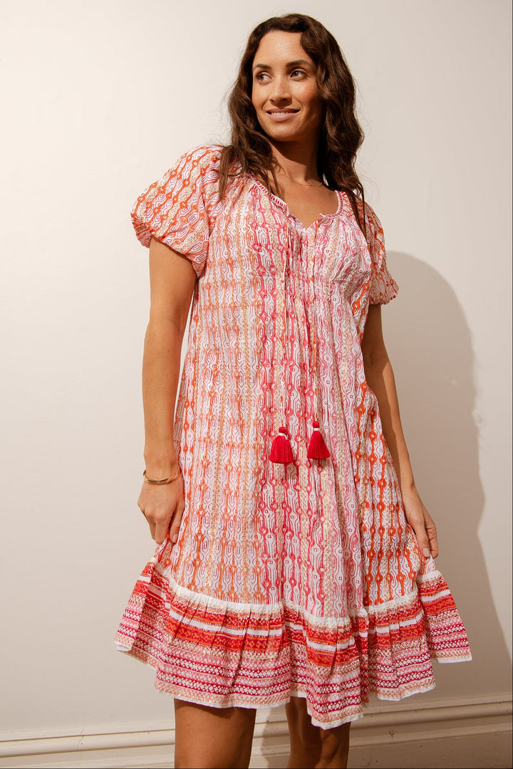 A woman wearing a knee-length orange sundress with a v-neckline, short sleeves, and a ruffle hemline. The dress features white geometric patterns and tassel details.