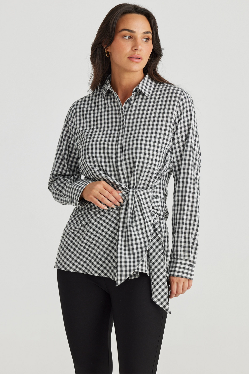 A woman wearing a black and white gingham shirt with a collar, buttons through the front, and an asymmetrical gathered waist tie.
