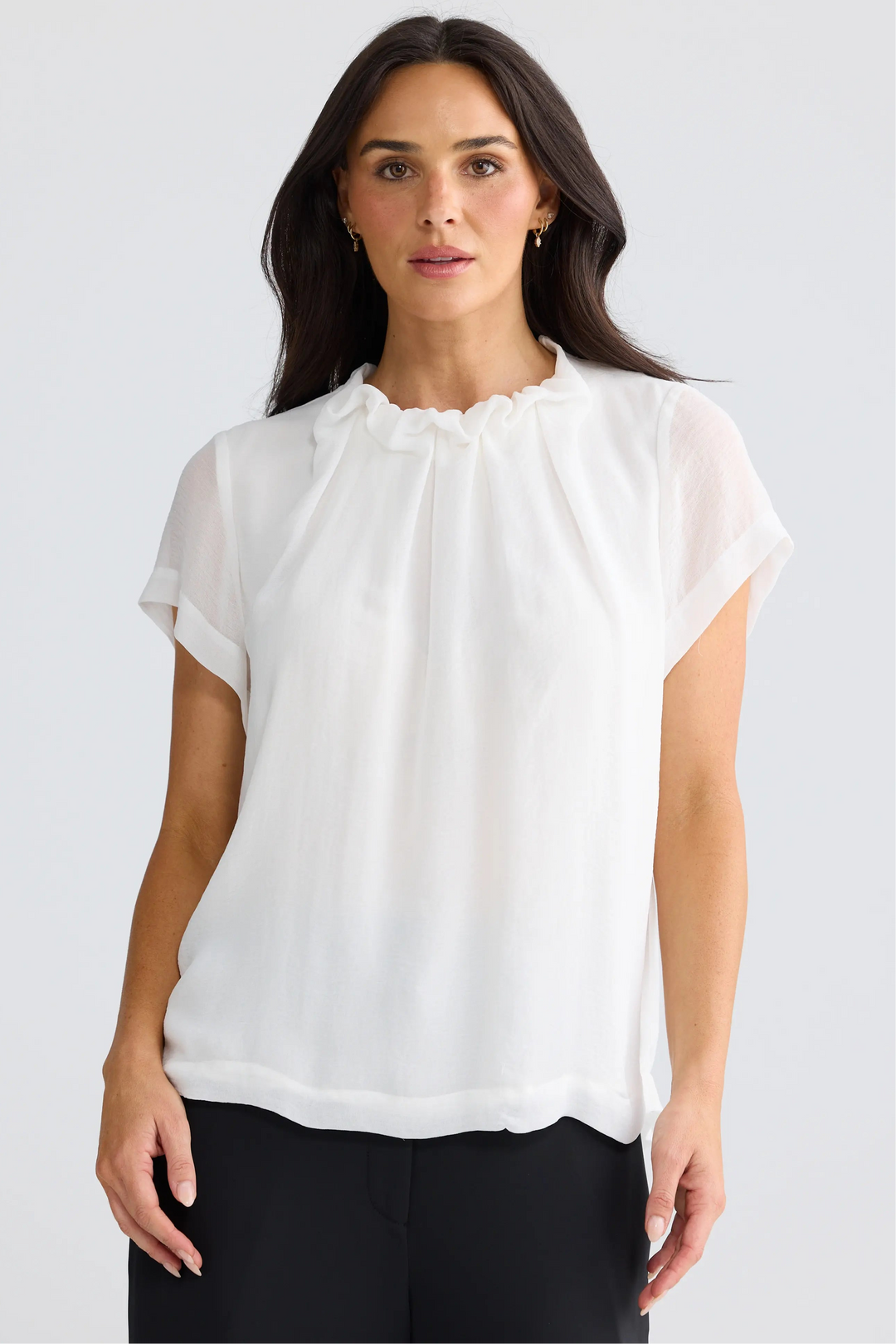 A woman wearing a white short-sleeve blouse with a ruffled high neckline, paired with black pants, photographed against a light gray studio background.
