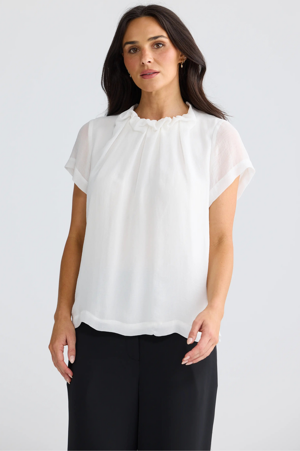 A woman modeling a white short-sleeve blouse with a ruffled high neckline, paired with black trousers, against a plain light gray studio background.