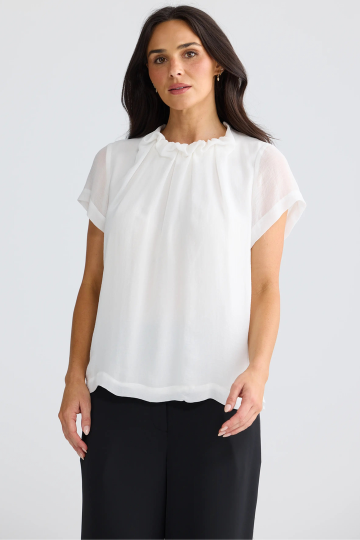 A woman modeling a white short-sleeve blouse with a ruffled high neckline, paired with black trousers, against a plain light gray studio background.