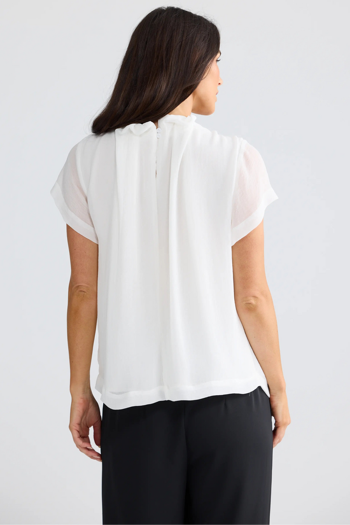 A woman viewed from the back wearing a white short-sleeve blouse with a gathered, pleated back detail and black pants, set against a plain light gray studio background.