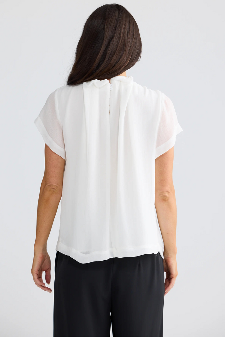 A woman viewed from the back wearing a white short-sleeve blouse with a gathered, pleated back and black pants, set against a plain light gray studio background.