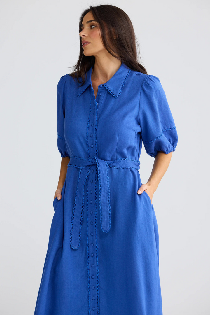 A woman wearing a bright blue midi dress with puff sleeves, a pointed collar, and a matching tie belt, photographed against a plain light gray studio background.