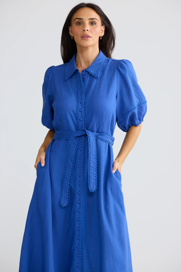 A woman modeling a bright blue midi dress with puff sleeves, a pointed collar, and a matching waist tie, set against a plain light gray studio background.