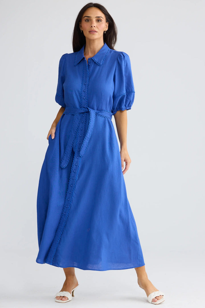 A woman modeling a bright blue midi dress with puff sleeves, a pointed collar, and a matching fabric tie belt, photographed against a light gray studio background.