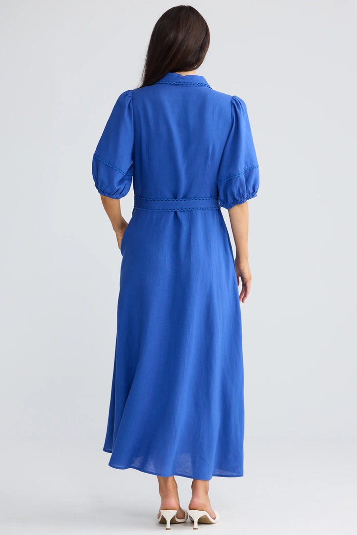 A woman viewed from the back wearing a bright royal blue midi dress with puff sleeves and a tie waist, paired with white heeled sandals, against a light gray studio background.
