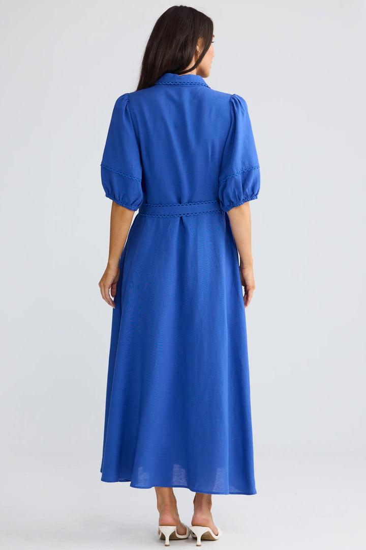 A woman viewed from the back wearing a bright royal blue midi dress with puff sleeves and a matching waist tie, paired with white heeled sandals, against a light gray studio background.