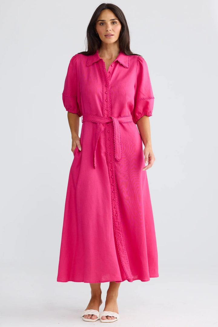 A woman modeling a bright pink midi shirt dress with short puff sleeves, a button-up front, and a matching tie belt, standing against a light gray studio background.