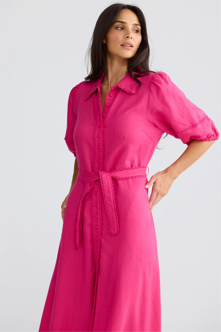A woman modeling a bright pink midi shirt dress with puff sleeves, a button-up front, and a matching tie belt, set against a plain light gray studio background.