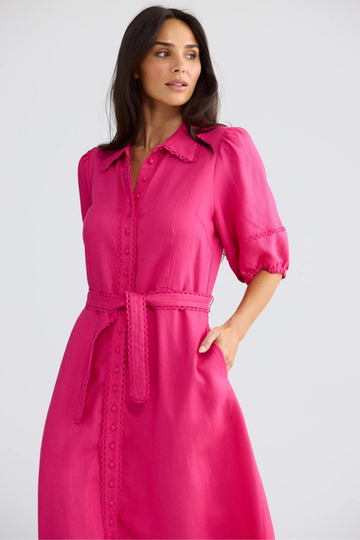 A woman wearing a bright pink midi shirt dress with puff sleeves, a matching tie belt, and scalloped edges, photographed against a light gray studio background.