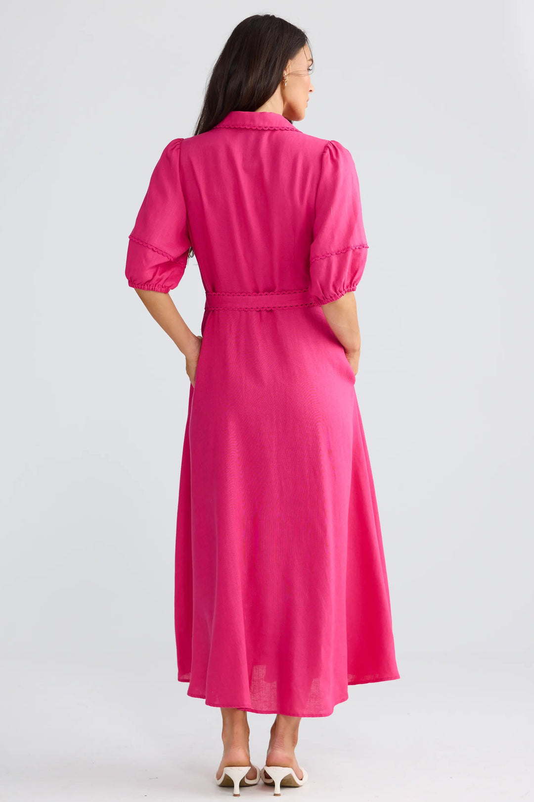 A woman is shown from the back wearing a bright pink midi dress with puff sleeves and a matching tie belt, standing against a light gray studio background.
