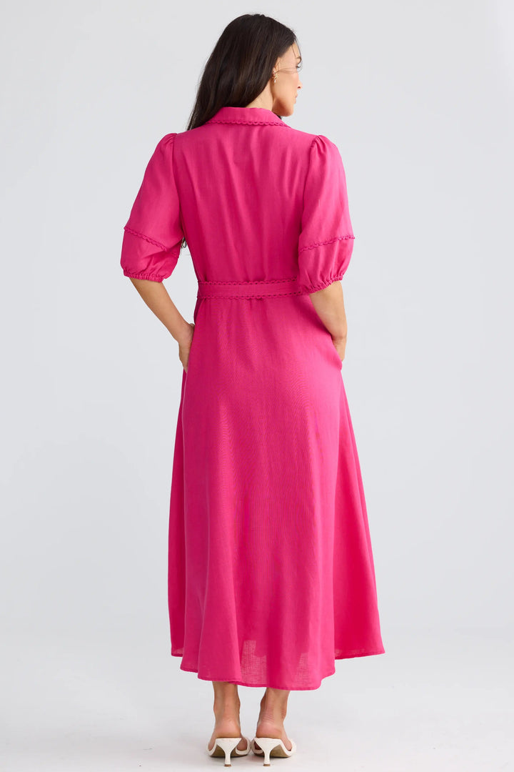 A woman is shown from the back wearing a bright pink midi dress with puff sleeves and a matching tie belt, standing against a light gray studio background.