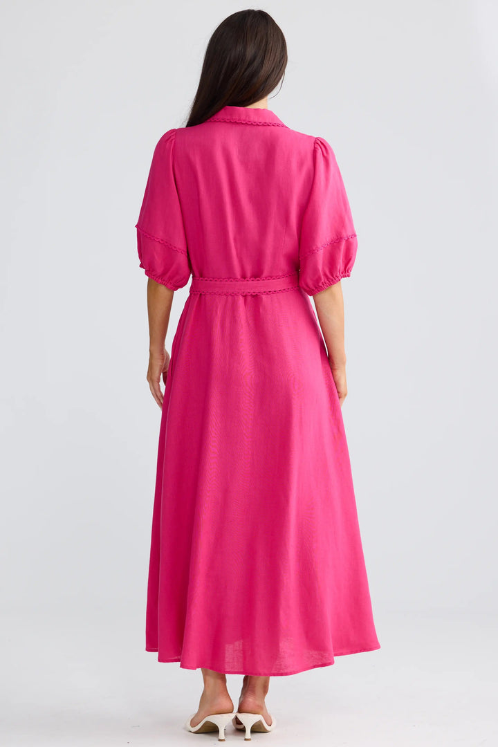 A woman viewed from the back wearing a bright pink midi dress with puff sleeves and a matching tie belt, standing against a plain light gray studio background.