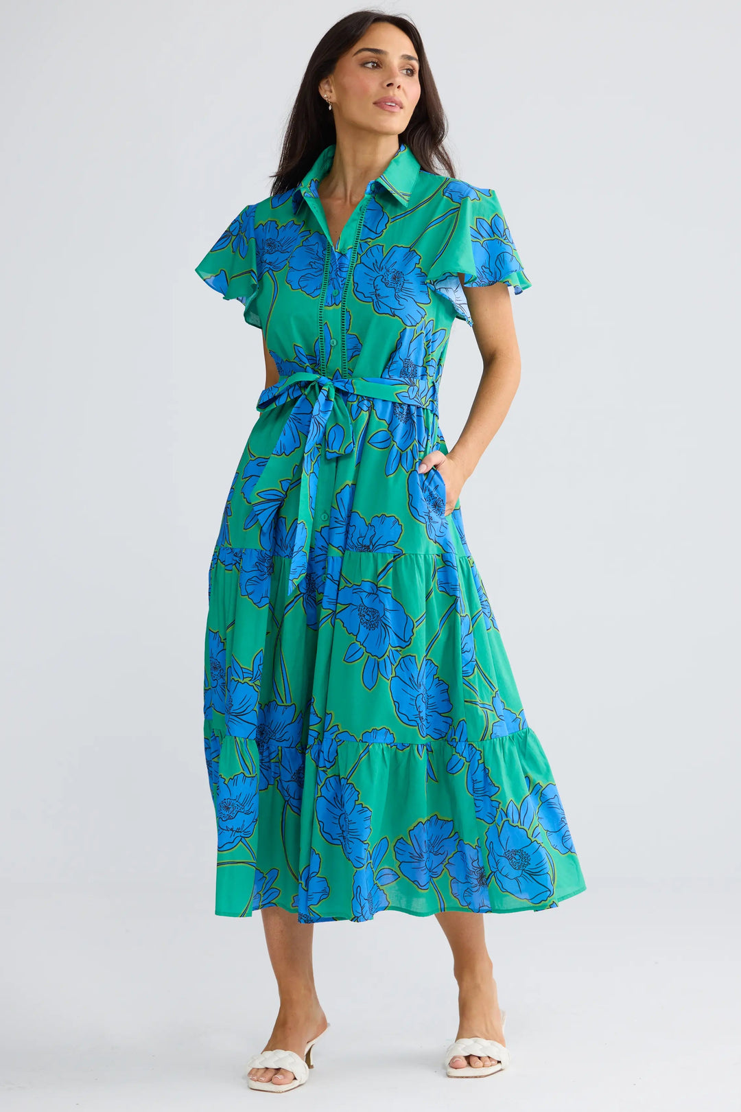 A woman modeling a green and blue floral print midi dress with short flutter sleeves, a collared neckline, and a matching tie belt, set against a plain light gray studio background.