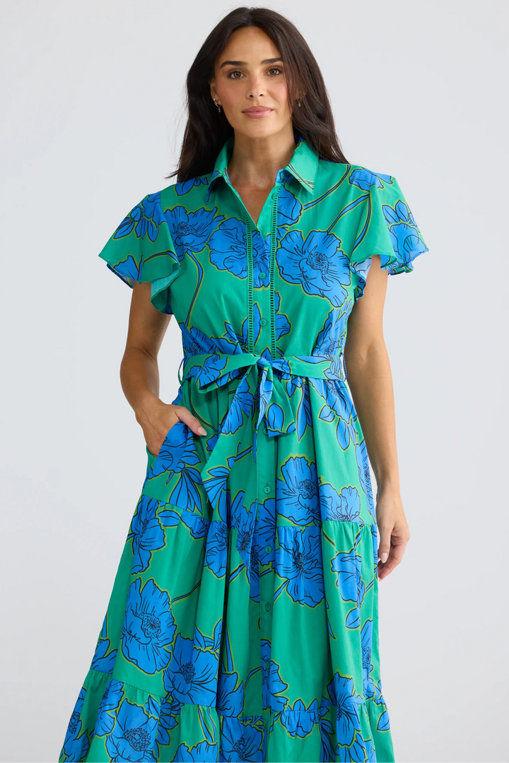 A woman modeling a green and blue floral print midi dress with a tie waist and short flutter sleeves, set against a plain light gray studio background.