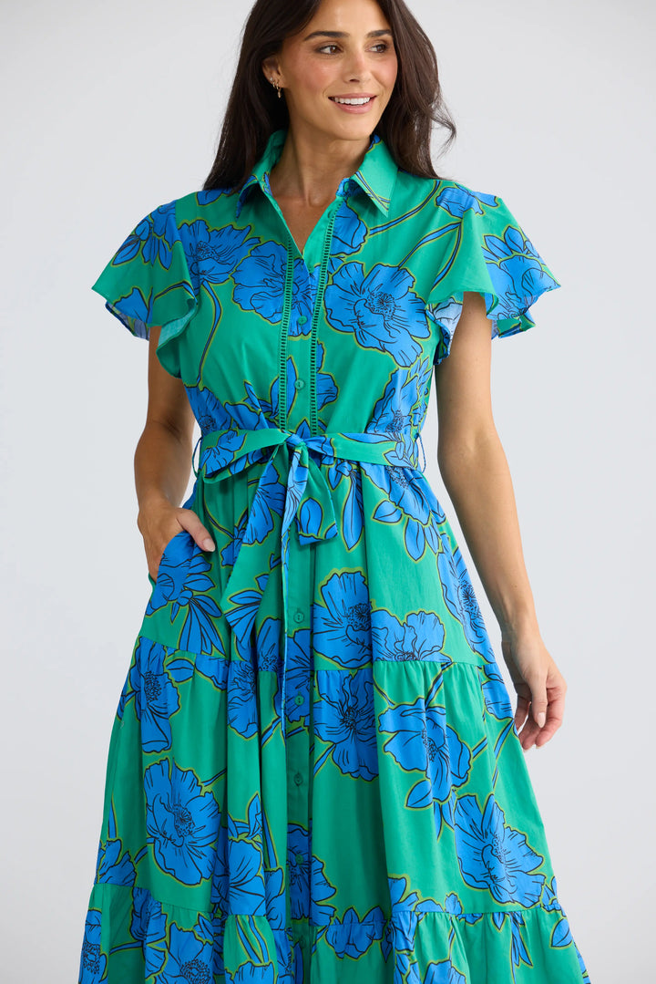A woman wearing a green and blue floral print midi dress with a tie waist and ruffled sleeves, photographed against a light gray studio background.