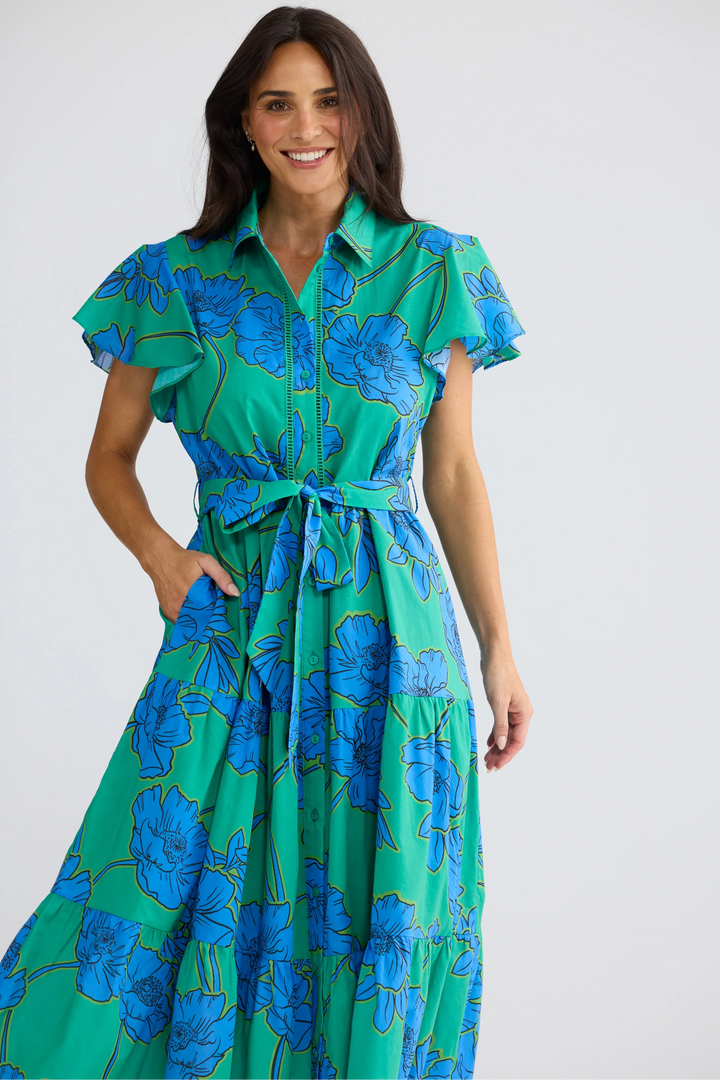 A woman modeling a green and blue floral print midi dress with short flutter sleeves, a collared neckline, and a matching tie belt at the waist, set against a plain light gray studio background.