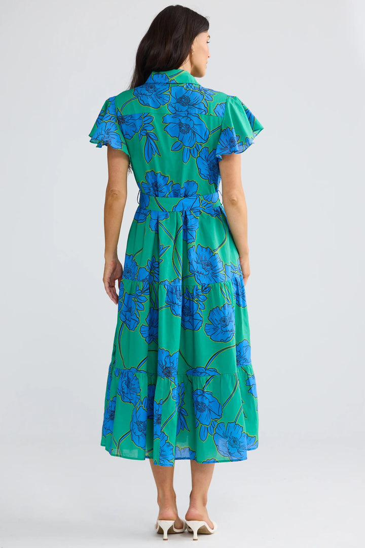 A woman viewed from the back wearing a green midi dress with a blue floral print, ruffled short sleeves, and a tie waist, paired with white heeled sandals against a light gray studio background.