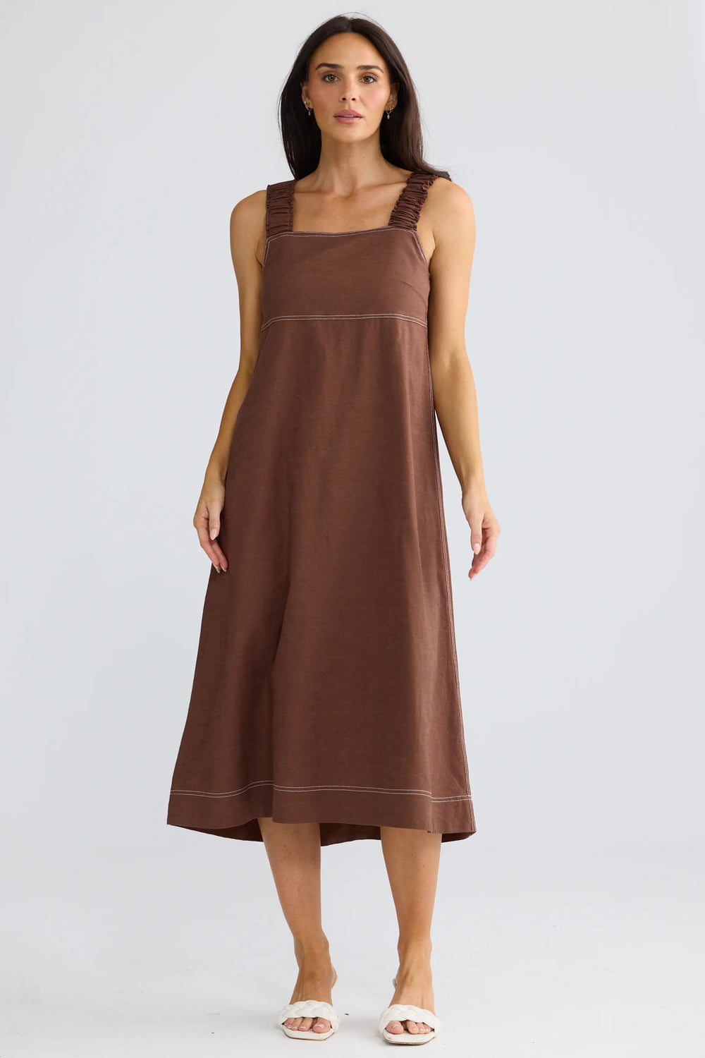 A woman modeling a sleeveless brown midi dress with a square neckline and wide shoulder straps, standing against a light gray studio background.