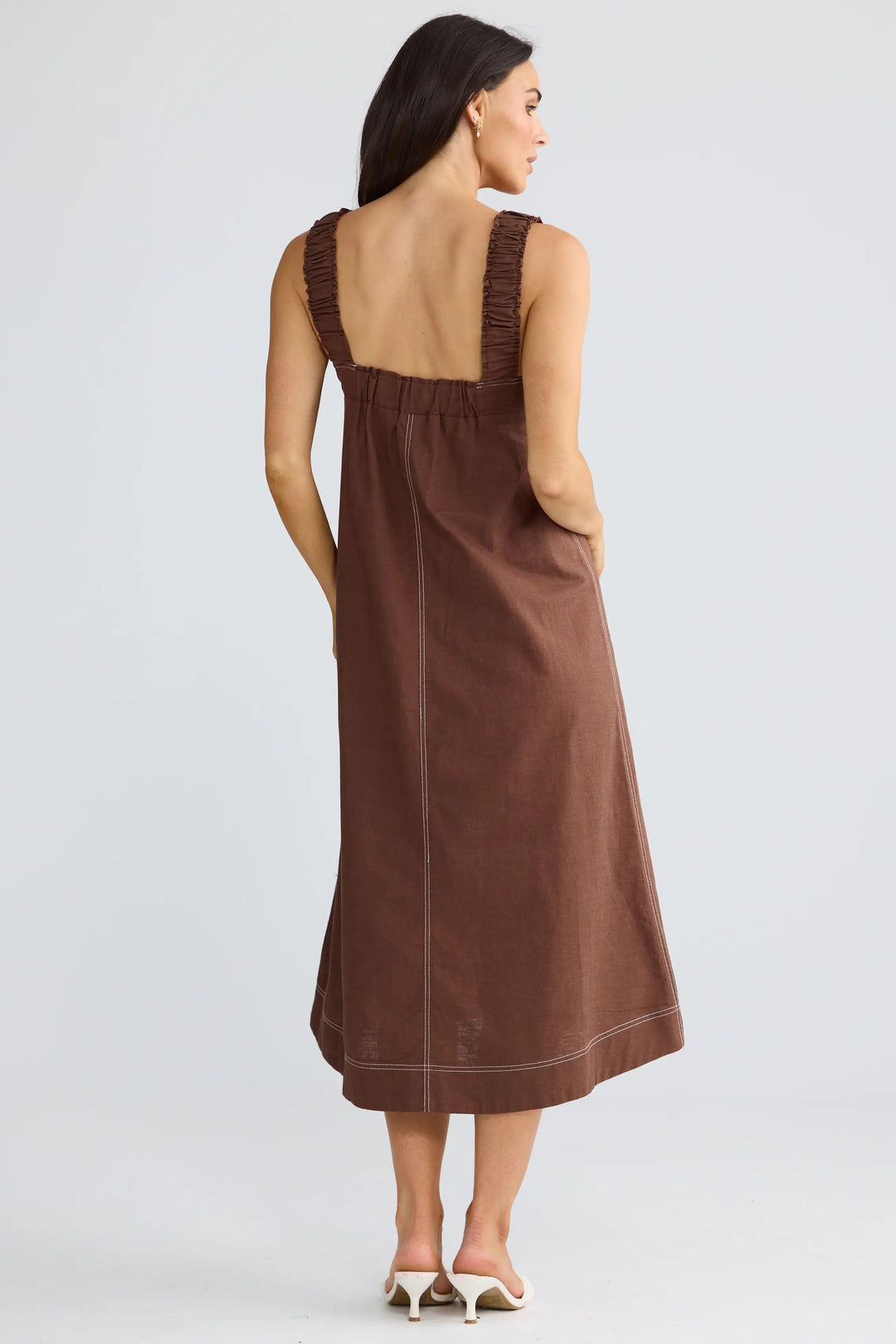 A woman wearing a brown sleeveless midi dress with a wide back and side pockets, shown from behind against a light gray studio background.