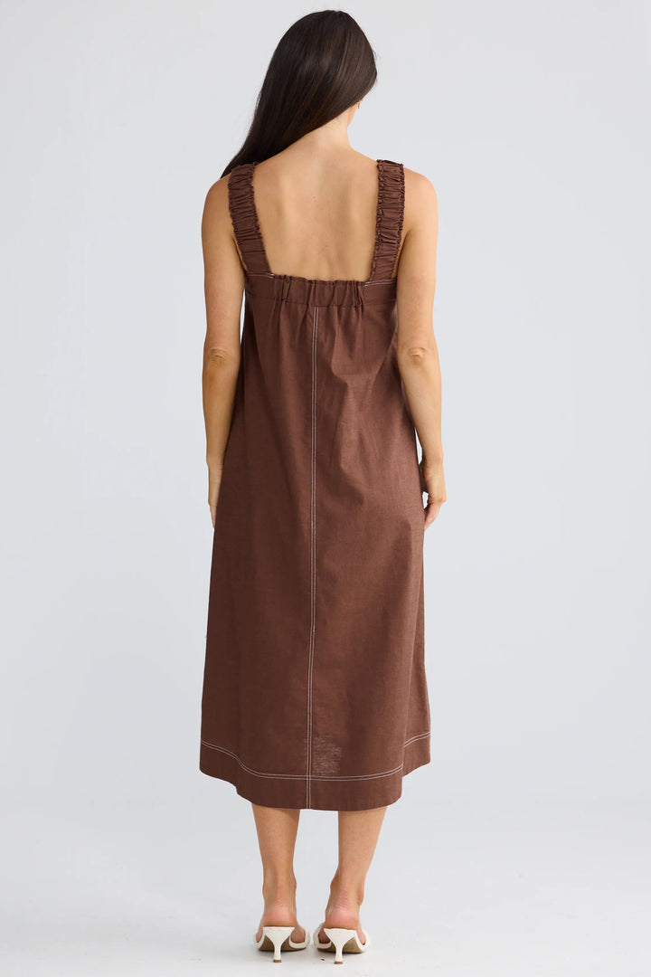 A woman viewed from the back wearing a sleeveless brown midi dress with wide shoulder straps and a gathered neckline, standing against a light gray studio background.