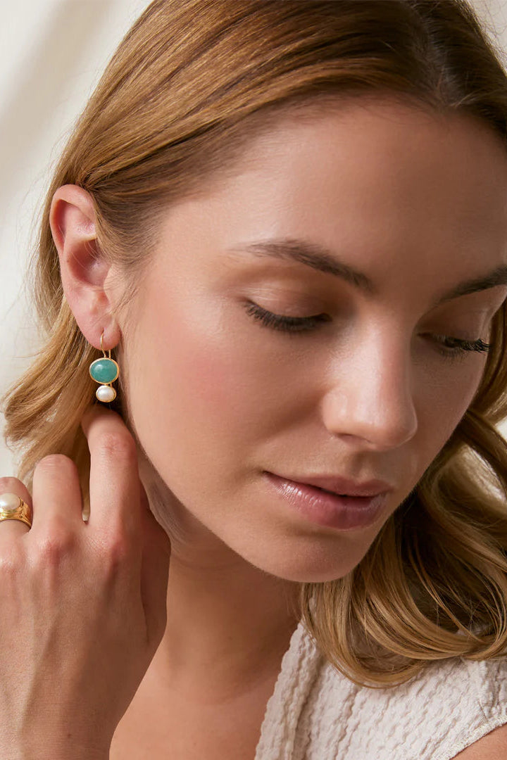 A close-up lifestyle shot of a woman wearing a gold hoop earring with a small round turquoise stone and a white bead drop, paired with a gold ring on her finger, set against a soft cream-colored draped background.