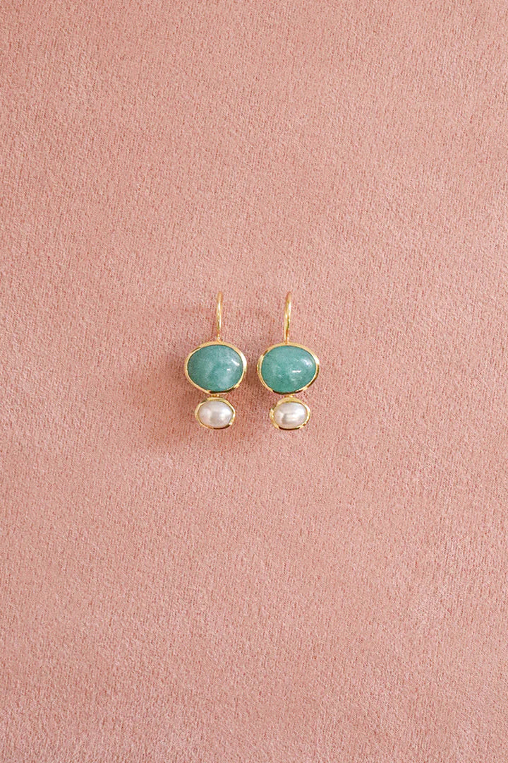 A pair of gold-tone drop earrings featuring oval light green cabochon stones and small white pearl-like drops, displayed on a textured dusty pink background.