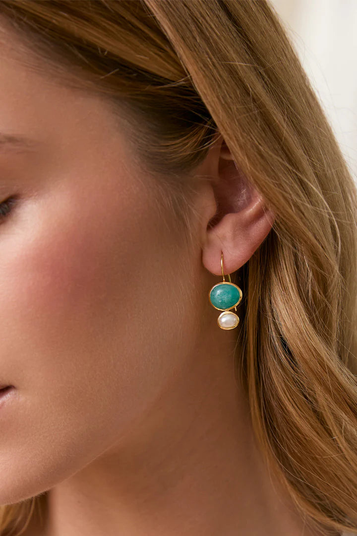 A close-up of a woman's ear wearing a gold dangle earring with a round turquoise-blue stone and a small white pearl.