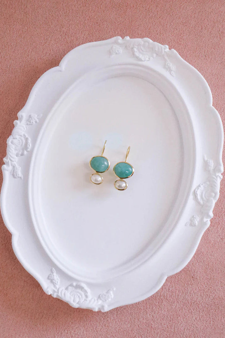 A pair of gold drop earrings with round turquoise-blue beads and small pearl-like drops, displayed on an ornate white oval plate against a dusty pink background.