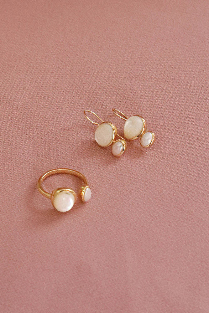 A pair of gold-tone drop earrings and a matching ring, each featuring a large round white pearl-like stone with smaller pearl-like accents, displayed on a dusty pink background.