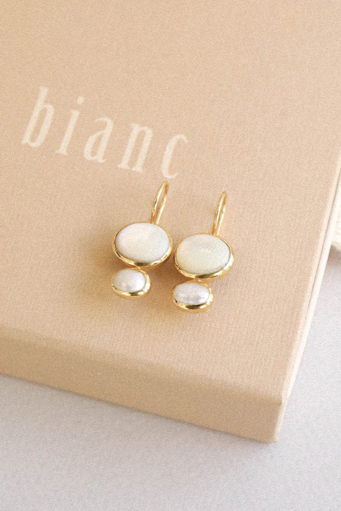 A pair of gold-tone drop earrings with oval white mother-of-pearl studs, displayed on a beige jewelry box printed with the brand name 'bianc'.