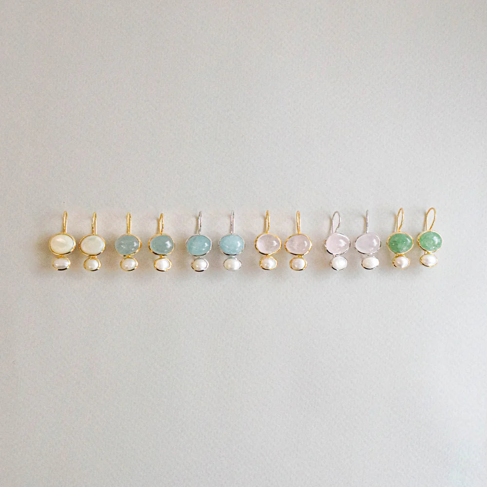 A row of small gold-tone earrings with oval gemstones in white, light blue, pink, and green, each hanging from a pearl bead, displayed on a light gray background.