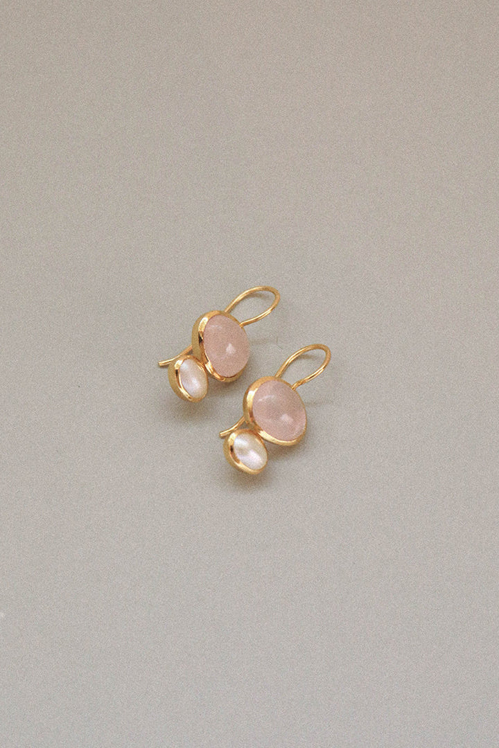 A pair of gold drop earrings featuring oval light pink stones and small teardrop-shaped white pearls, displayed on a neutral gray background.