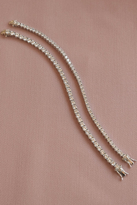 Two silver tennis bracelets with clear crystals, displayed on a dusty pink textured fabric background.
