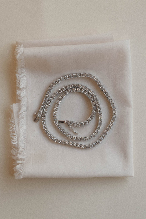 A silver beaded necklace with a small charm, coiled on a folded white fabric napkin against a light beige background.