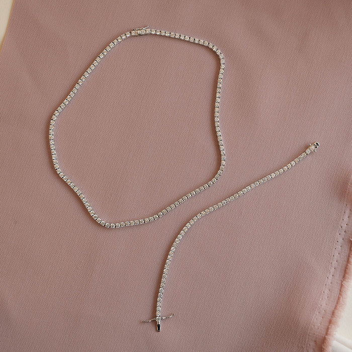 A delicate silver tennis-style necklace with clear rhinestones, arranged in a loose loop on a soft dusty pink fabric background.