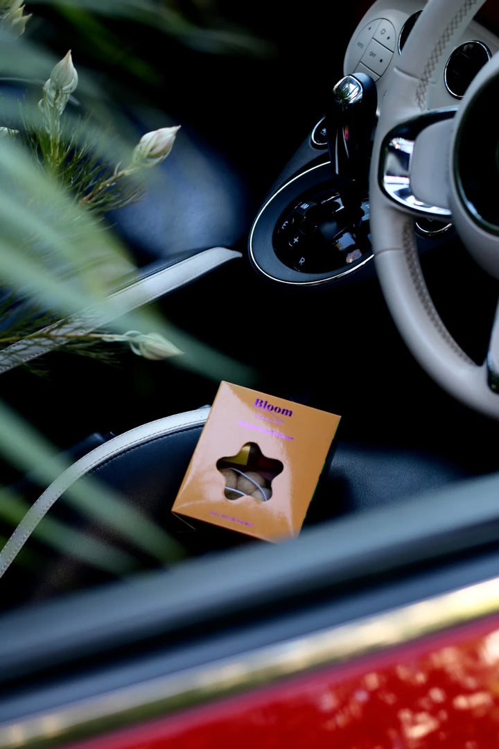 Flower shaped felt air freshener packaging on a car seat with steering wheel and cup holder in the background.