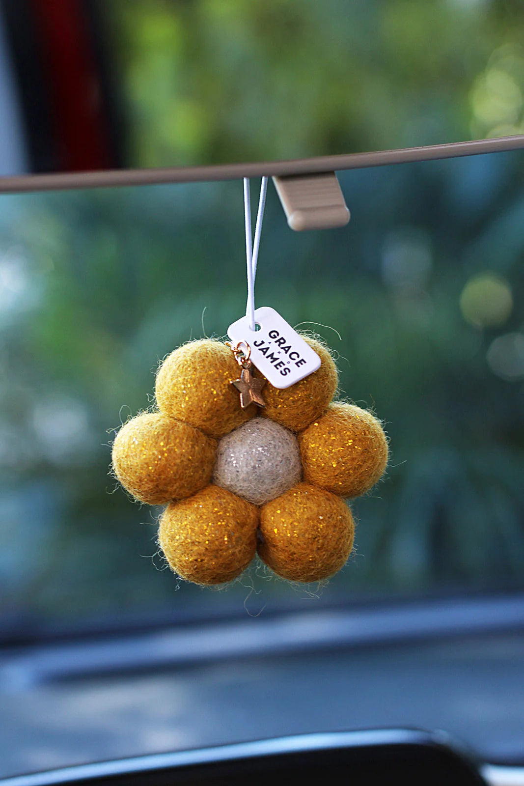 Yellow flower-shaped felt air freshener hanging from a car's rearview mirror with a blurred green background.