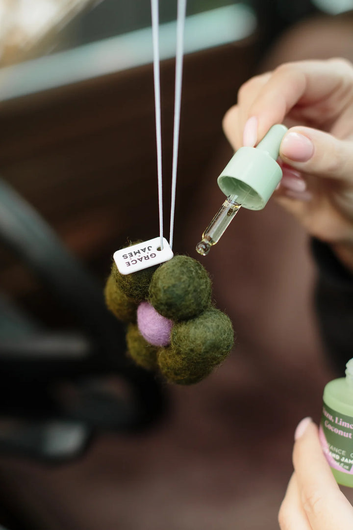 Car air freshener with felt balls and a dropper bottle held by a hand. Yuzu Lime & Coconut