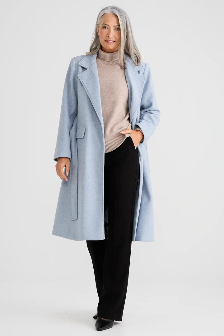 Woman wearing a light blue coat over a beige sweater and black pants on a white background