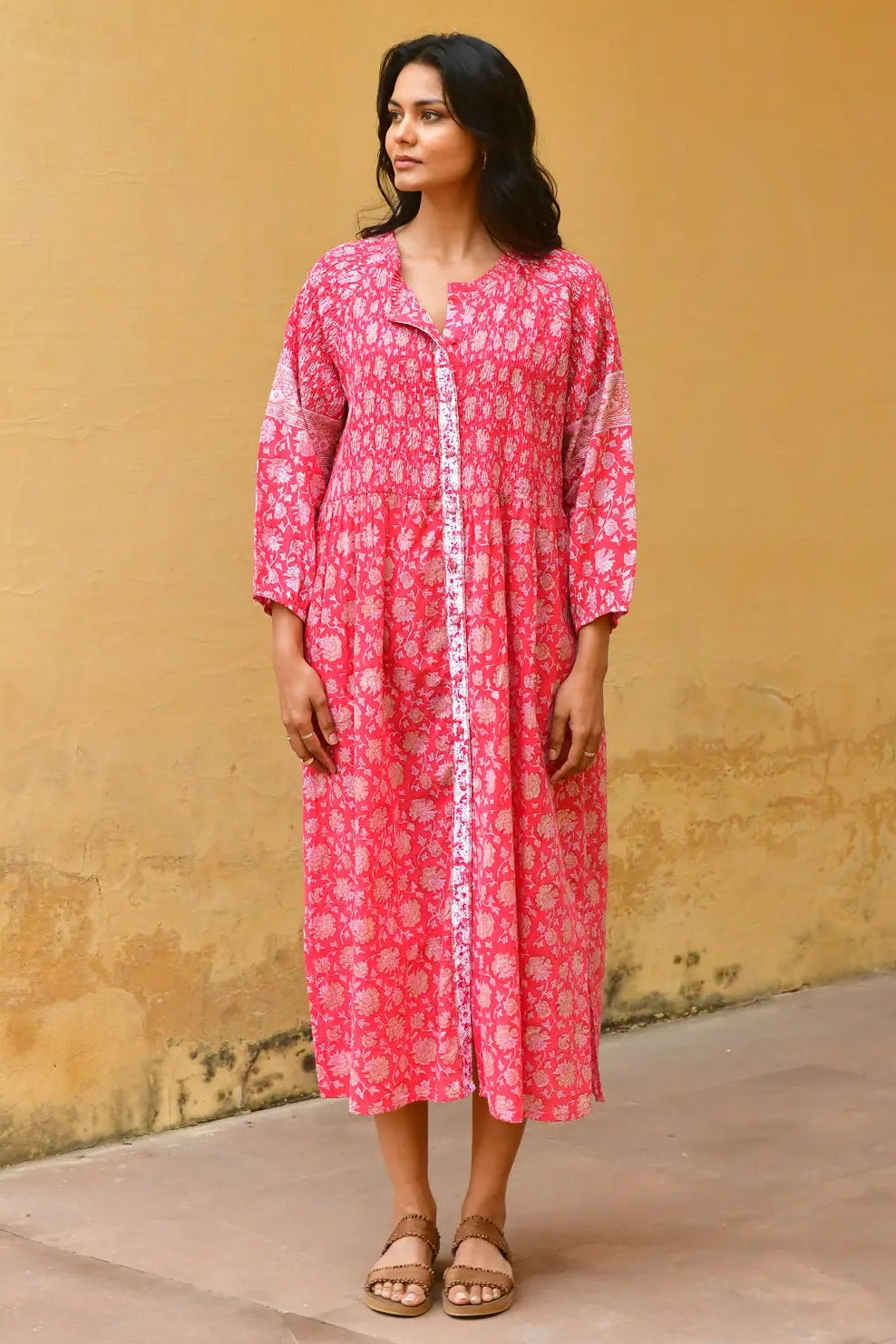 Woman wearing a pink dress with white patterns against a yellow wall. Azure & Indigo Camillia Maxi Dress Cherry.