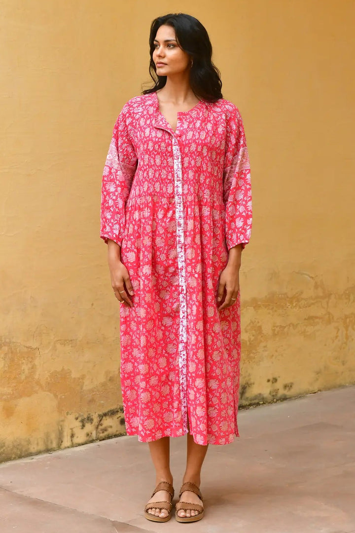 Woman wearing a pink dress with white patterns against a yellow wall. Azure & Indigo Camillia Maxi Dress Cherry.