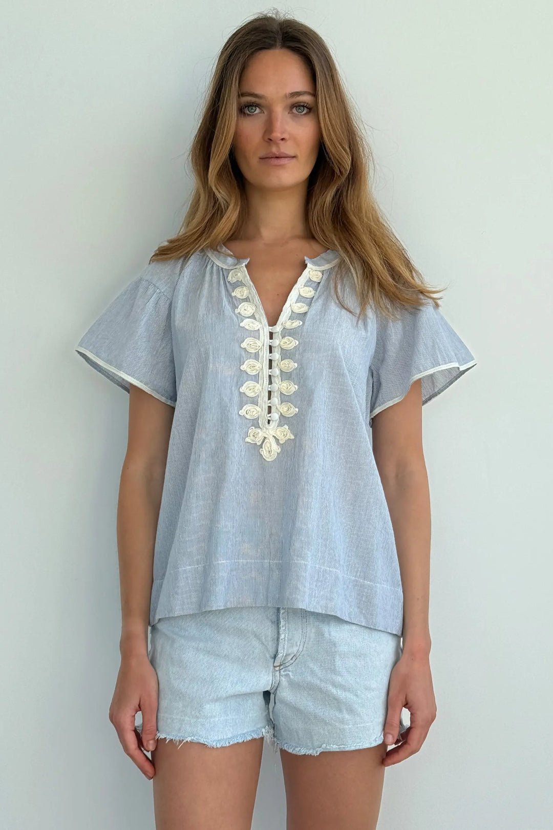 Woman wearing a light blue embroidered top and denim shorts against a white background
