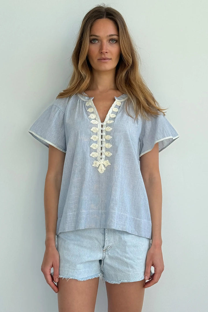 Woman wearing a light blue embroidered top and denim shorts against a white background