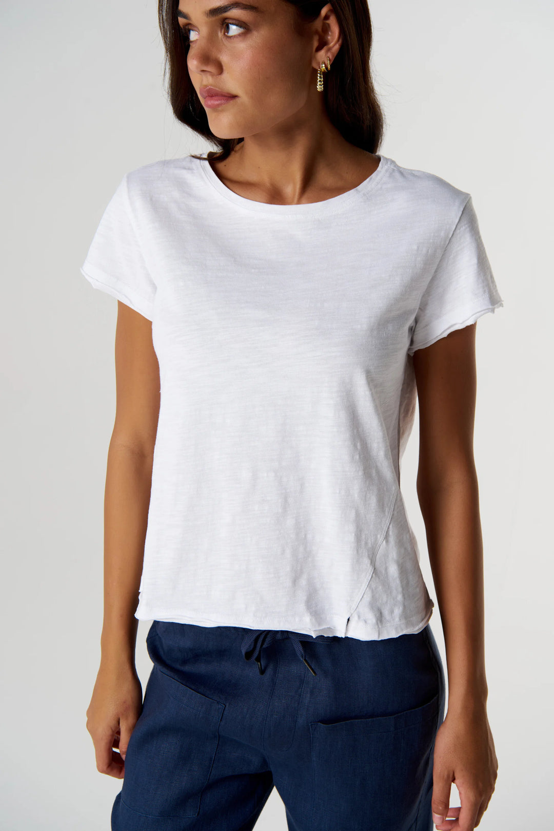 A woman wearing a white short-sleeve crewneck t-shirt and navy blue pants, photographed against a light gray studio background.