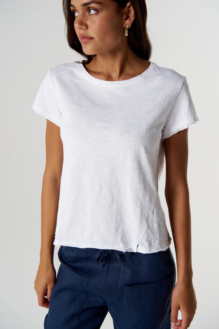 A woman wearing a white short-sleeve crewneck t-shirt and navy blue pants, photographed against a light gray studio background.