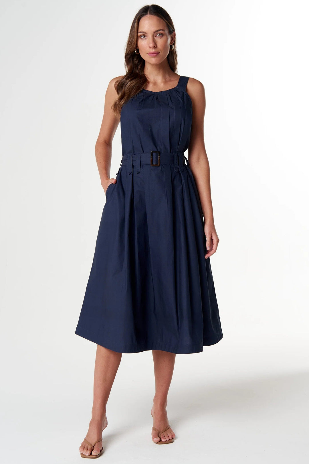 A woman modeling a sleeveless navy blue midi dress with a matching belt and buckle, standing against a plain white studio background.
