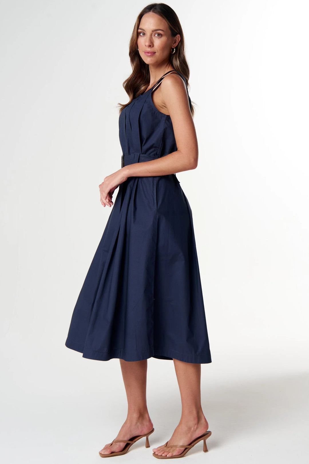 A woman modeling a sleeveless navy midi dress with thin straps, a gathered waist, and a flared skirt, paired with tan heeled sandals against a plain light gray studio background.