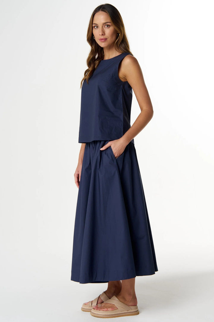 A woman modeling a navy blue sleeveless top and matching high-waisted maxi skirt set, paired with beige slide sandals, against a plain white studio background.